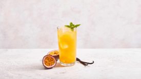A clear glass with an orange-colored passionfruit flavored beverage in front of a light pink background.