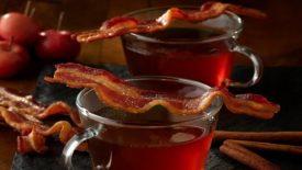 Two mugs of apple cider with bacon across the top of them
