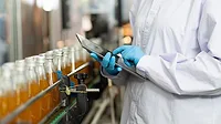 Person in lab coat and gloves working on a tablet A cropped image of a person wearing a white lab coat and blue gloves, looking at a tablet while standing in front of a line of bottles.
