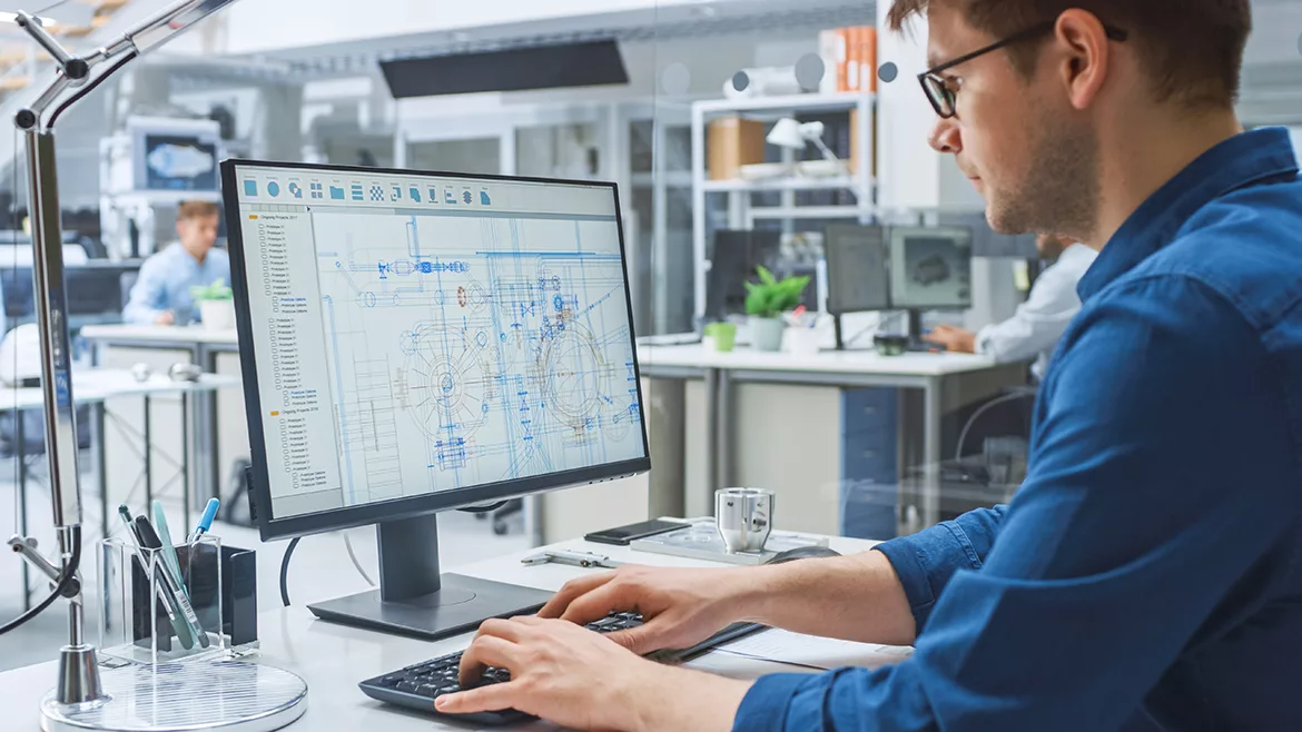 An engineer working on his desktop computer in an office and another engineer in the blurred background.