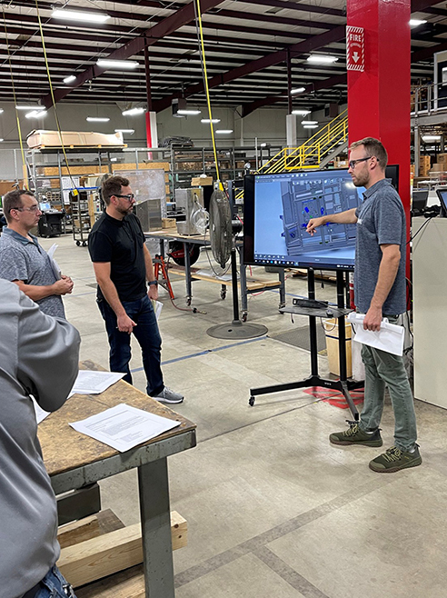 Three men examine a CAD drawing on a large screen in a warehouse, one pointing to the screen.