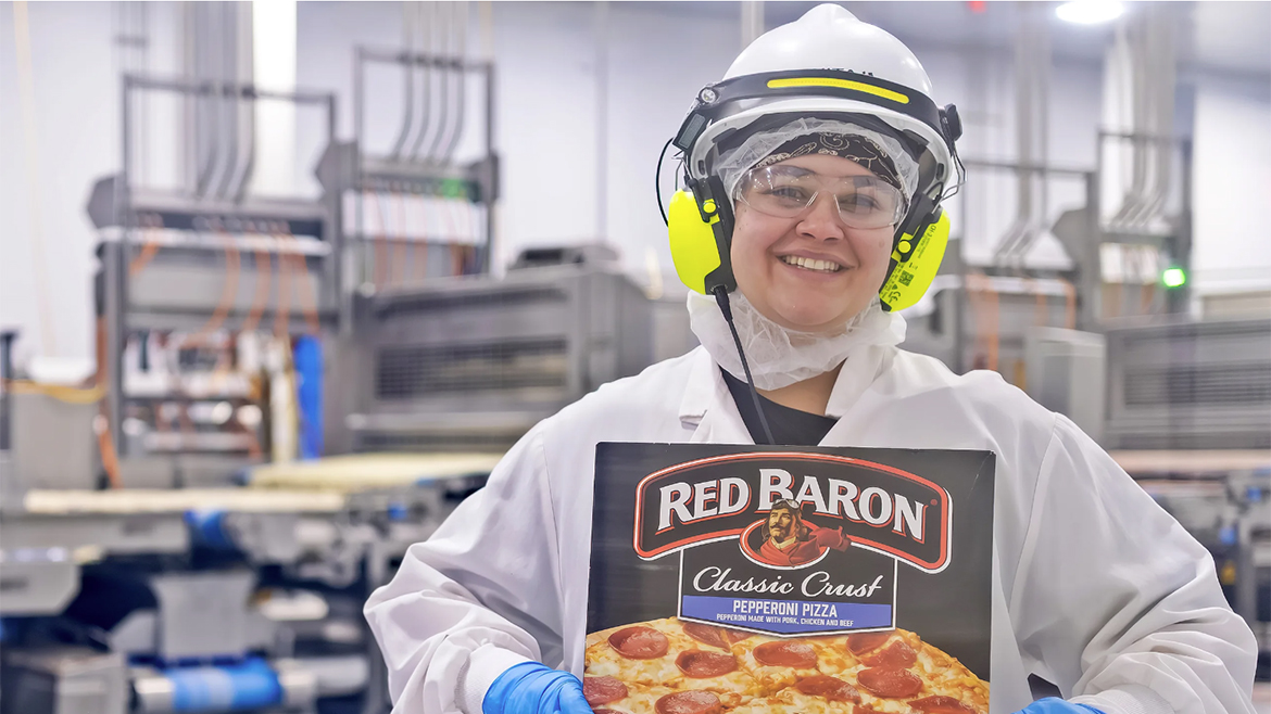 A smiling food production worker in safety gear holds a Red Baron Pepperoni Pizza box in a factory setting.
