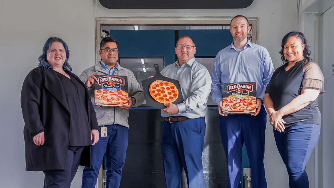 Five people with Red Baron pizzas at the manufacturing facility.