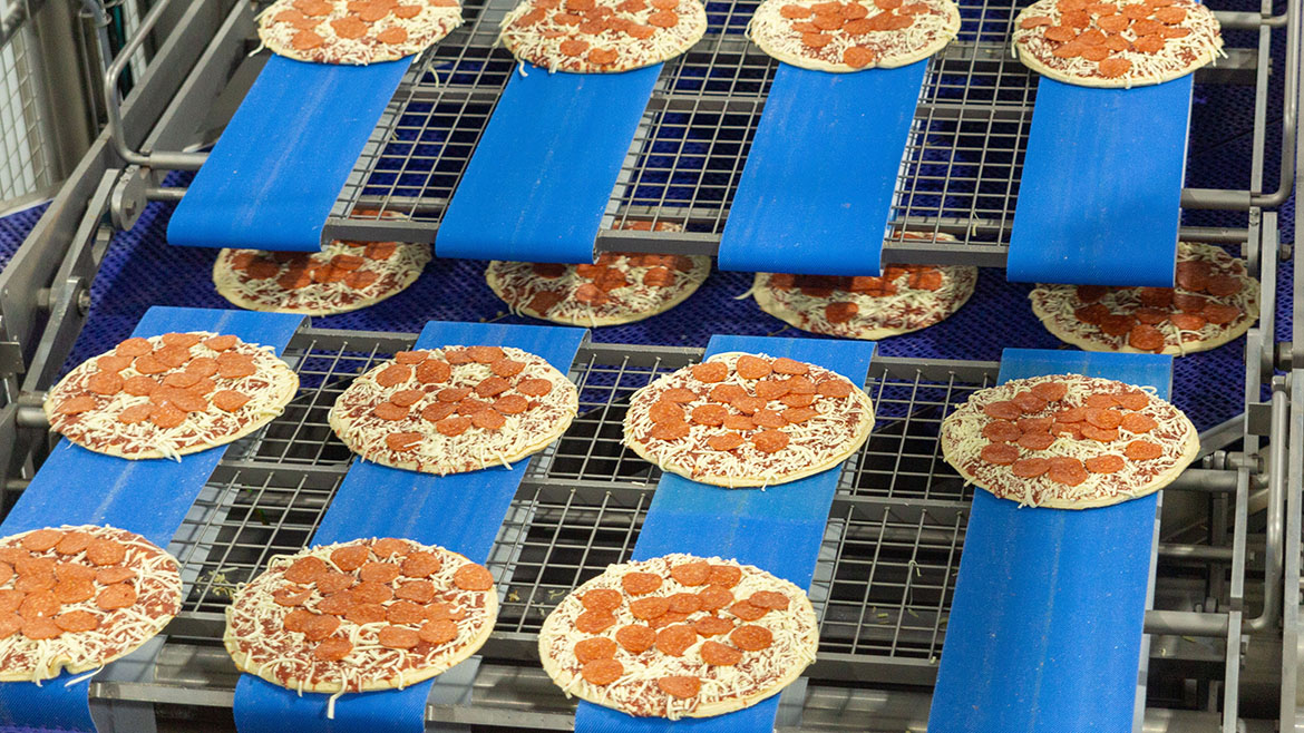 A line of uncooked pepperoni pizzas moving along blue conveyor belts in a food processing plant.