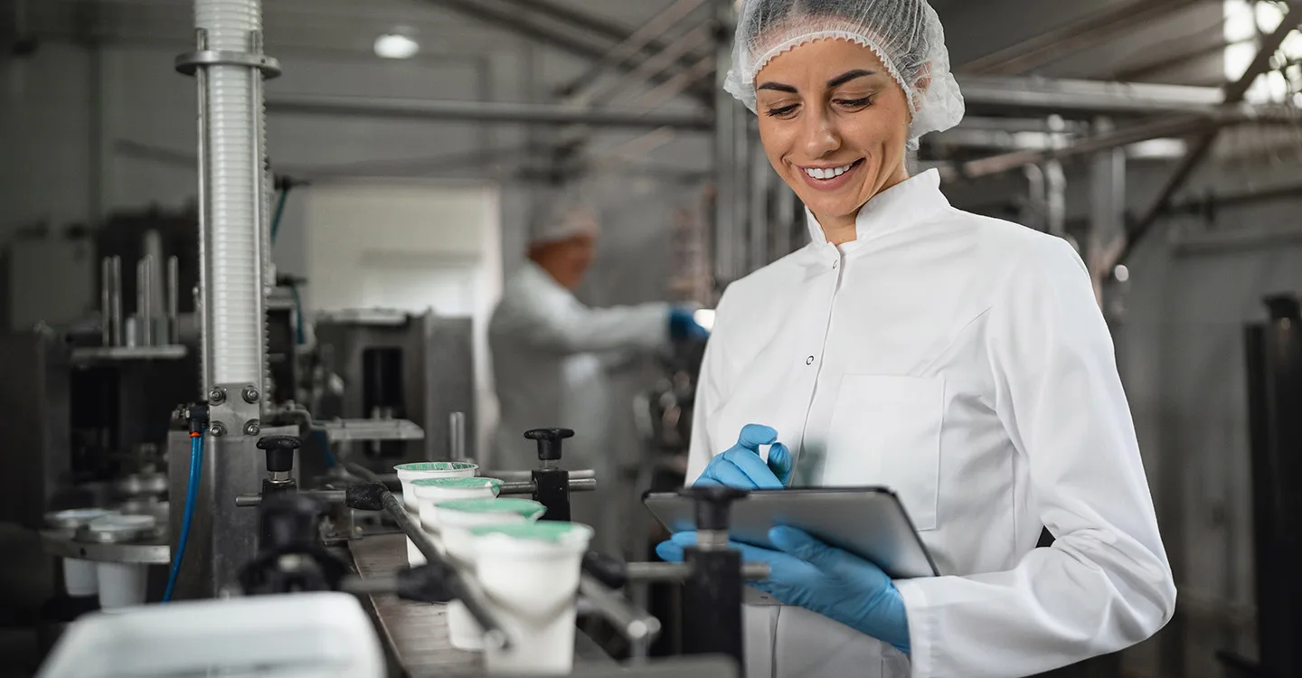 Technician inspects products in a dairy manufacturing assembly line. Another technician is shown in the blurred background.