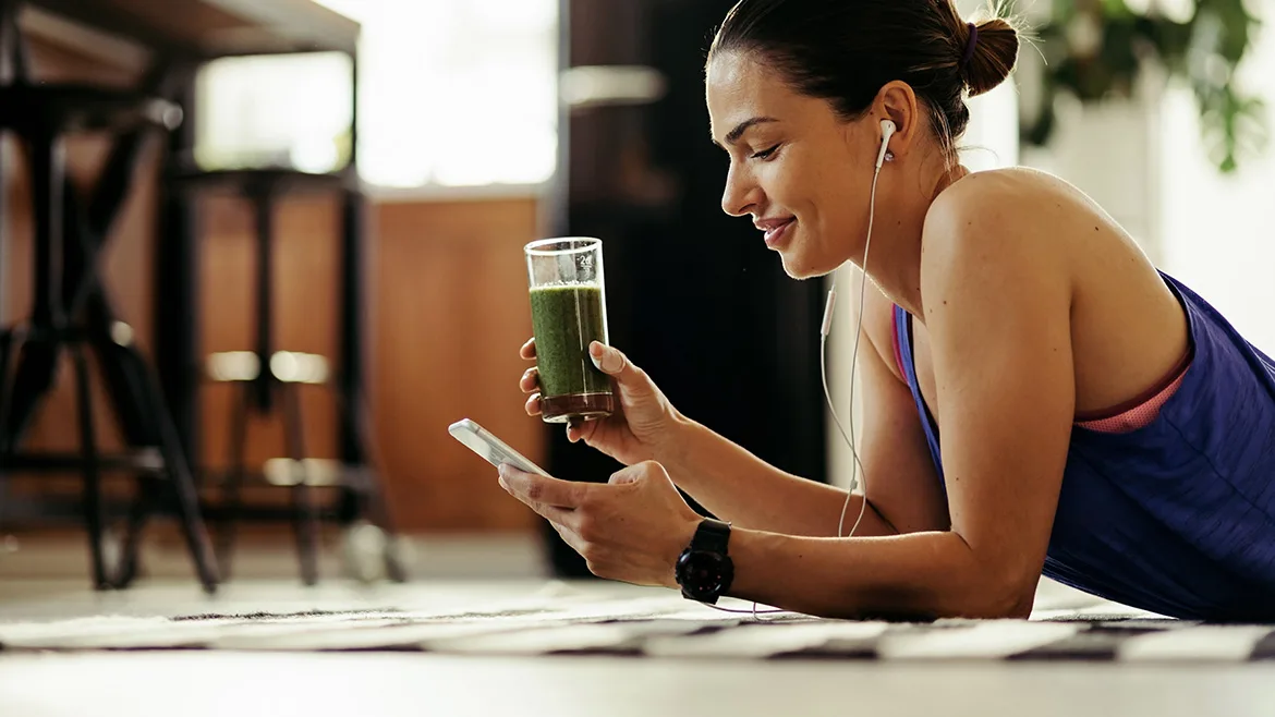 Woman with wired headphones is on her phone laying on her stomach on the floor and holding a green protein shake.
