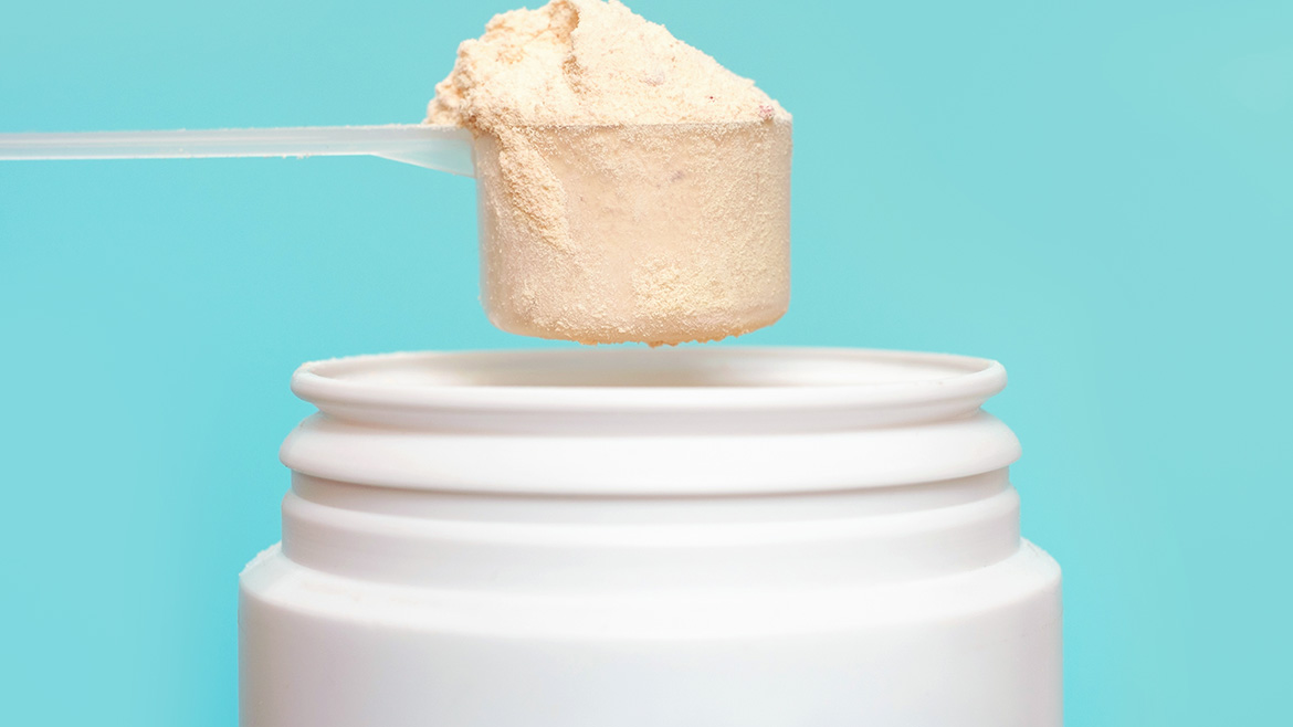 An overflowing scoop of protein powder being hold over a white container on a light blue background.