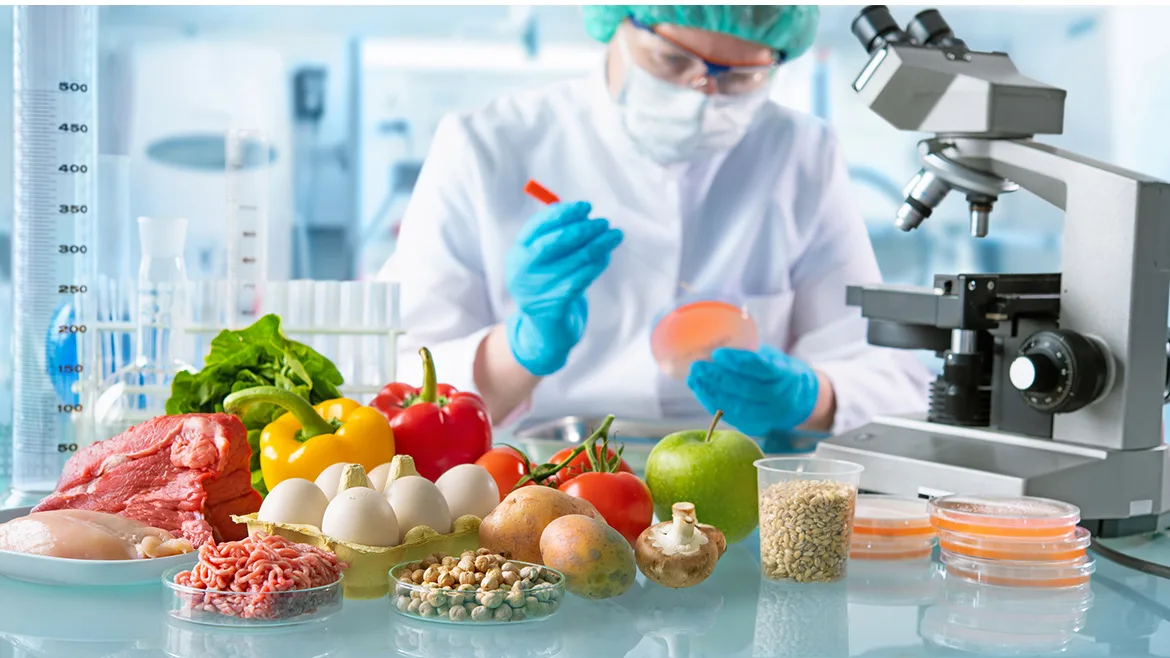 A food scientist in protective clothing and eye gear is working in a lab and examining petri dishes, fresh vegetables, fruits, seeds, eggs and meet in from of her.