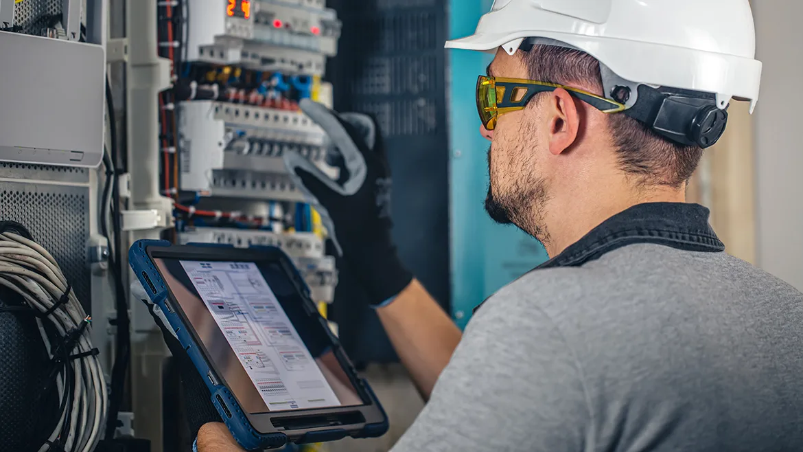 Worker in a white safety hat and glasses is using a digital tablet to examine equipment.