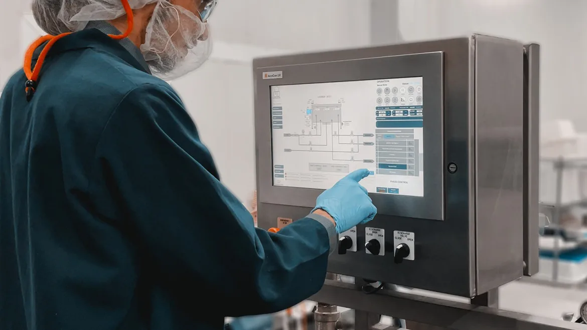 Blentech machine operator in safety gear, gloves, hair net, safety glasses and face covering, touching a monitor screen.