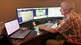 A man is working food safety monitoring on a desktop with three monitors. An open laptop is on the left side of the desk.