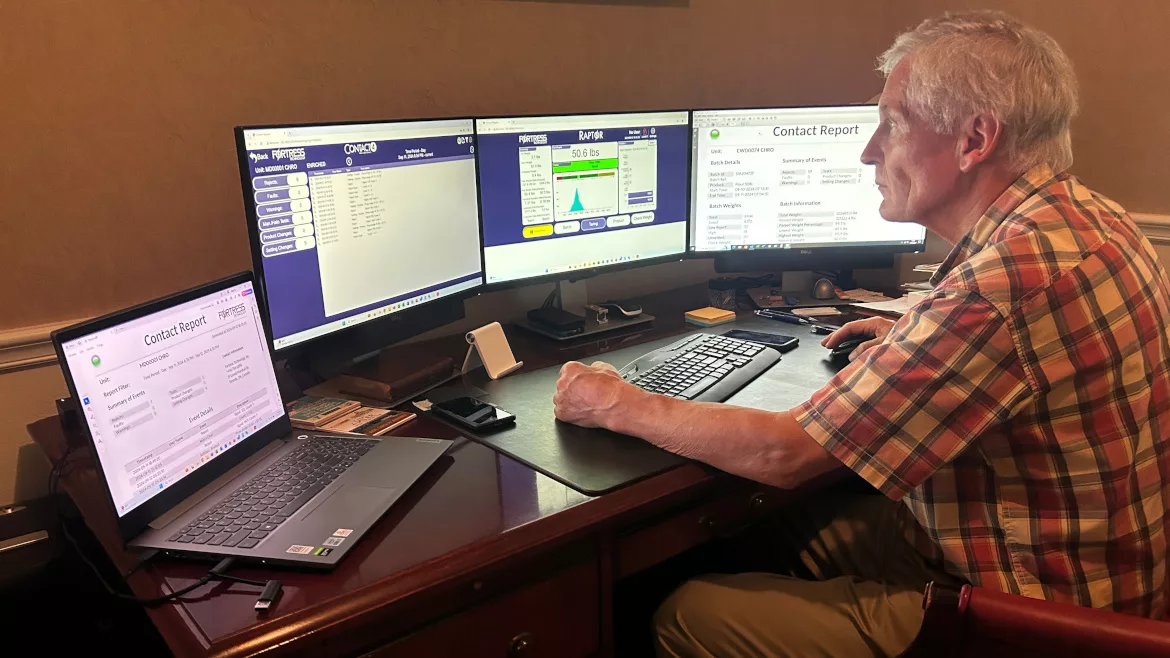 A man is working food safety monitoring on a desktop with three monitors. An open laptop is on the left side of the desk.