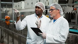 Two male manufacturing workers with tablet, wearing white coats, gloves and hairnets are pointing at something.