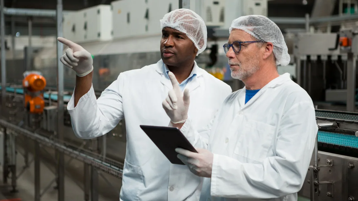 Two male manufacturing workers with tablet, wearing white coats, gloves and hairnets are pointing at something.
